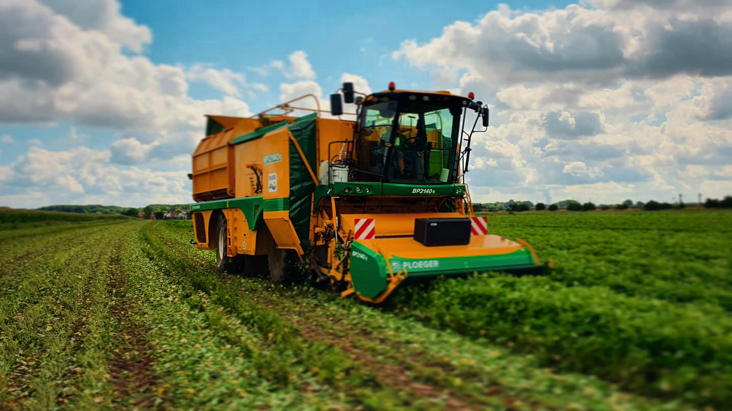 Eine große Erntemaschine fährt über ein grünes Feld, während dichter Himmel und weite Landschaft die Szene umrahmen.